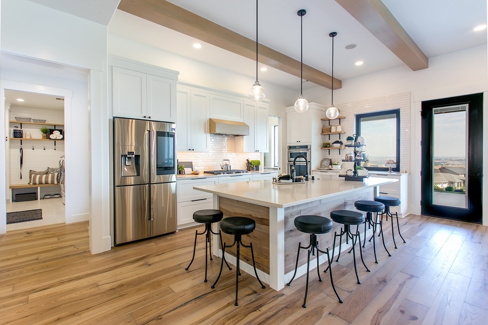 Inspiration for a country l-shaped kitchen in Seattle with shaker cabinets, white cabinets, white splashback, metro tiled splashback, stainless steel appliances, medium hardwood flooring, an island, brown floors and white worktops.