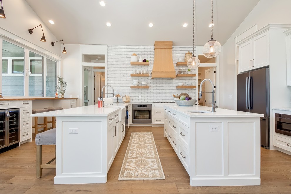 Inspiration for a large contemporary u-shaped open plan kitchen in Boise with a belfast sink, recessed-panel cabinets, white cabinets, composite countertops, white splashback, metro tiled splashback, stainless steel appliances, medium hardwood flooring, an island, brown floors and white worktops.