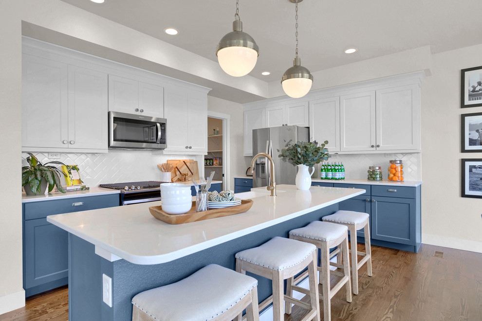 This is an example of a beach style kitchen in Denver with a belfast sink, blue cabinets, engineered stone countertops, white splashback, ceramic splashback, stainless steel appliances, an island, white worktops, shaker cabinets and medium hardwood flooring.