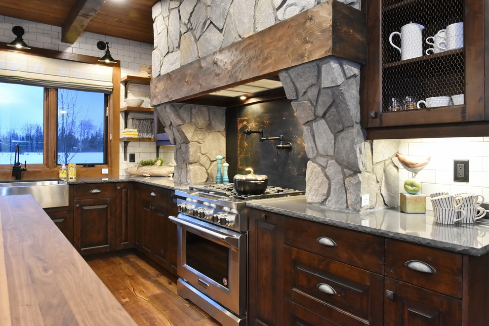 Bild på ett mellanstort rustikt brun brunt kök, med en rustik diskho, luckor med upphöjd panel, skåp i mörkt trä, träbänkskiva, vitt stänkskydd, stänkskydd i tunnelbanekakel, rostfria vitvaror, mellanmörkt trägolv, en köksö och brunt golv