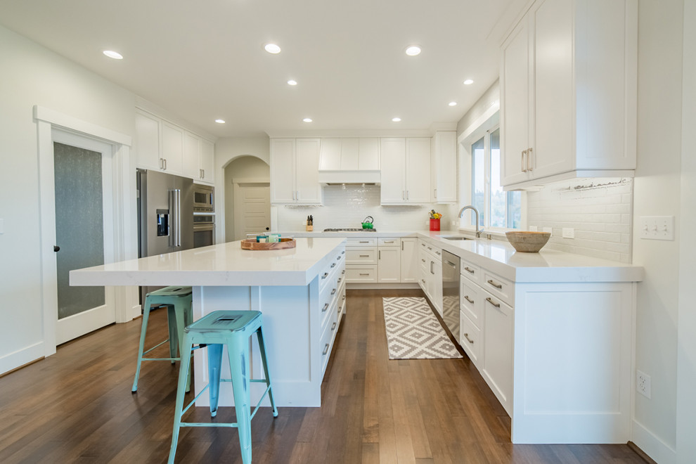 Medium sized classic u-shaped kitchen/diner in Seattle with a submerged sink, recessed-panel cabinets, white cabinets, engineered stone countertops, white splashback, metro tiled splashback, stainless steel appliances, medium hardwood flooring, an island, brown floors and white worktops.