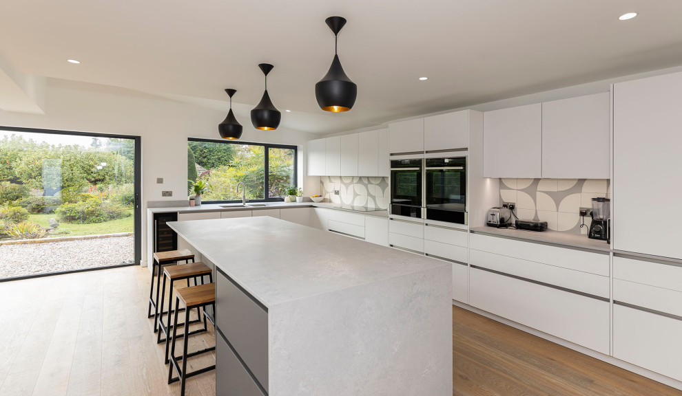 Photo of a large scandinavian open plan kitchen in Hertfordshire with a built-in sink, recessed-panel cabinets, granite worktops, mosaic tiled splashback, black appliances, laminate floors, an island, beige floors and grey worktops.