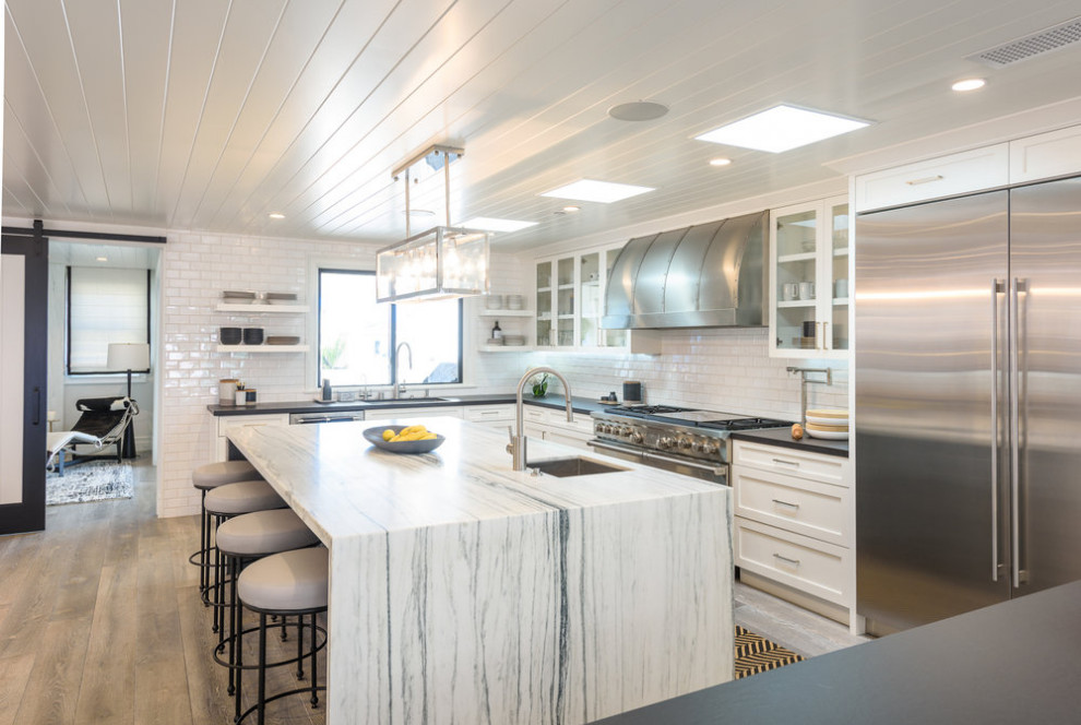 Large beach style u-shaped open plan kitchen in Los Angeles with a submerged sink, shaker cabinets, white cabinets, engineered stone countertops, white splashback, cement tile splashback, stainless steel appliances, medium hardwood flooring, an island, beige floors and white worktops.