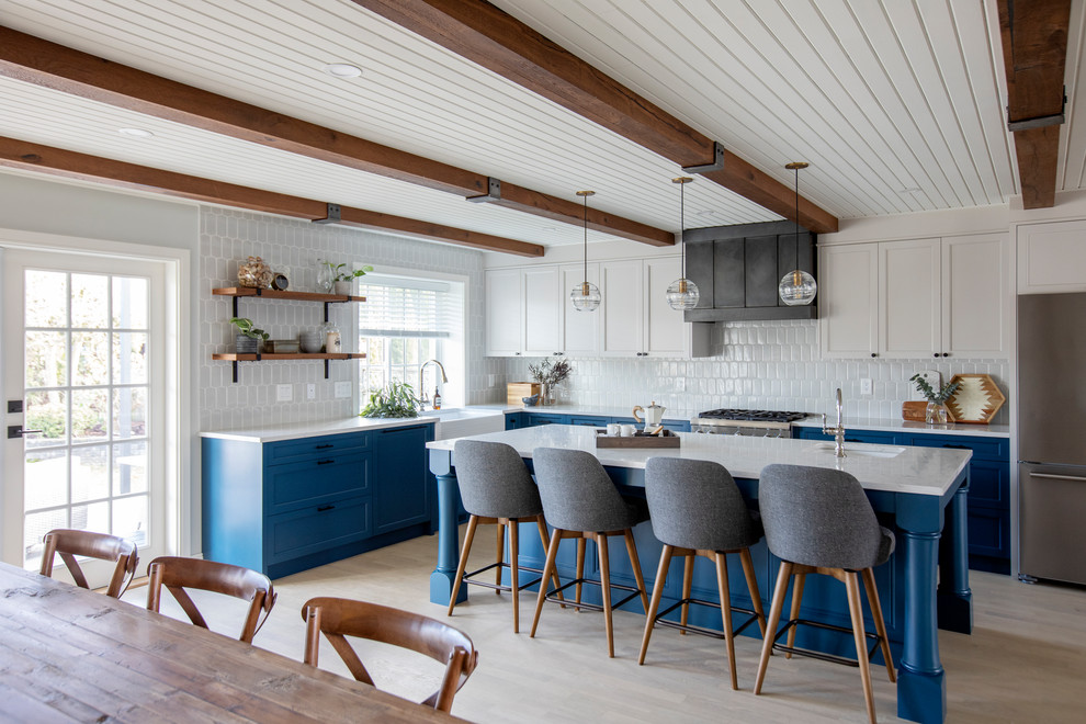 Bild på ett lantligt vit vitt kök, med skåp i shakerstil, bänkskiva i kvartsit, rostfria vitvaror, en köksö, en rustik diskho, blå skåp, vitt stänkskydd, ljust trägolv och beiget golv