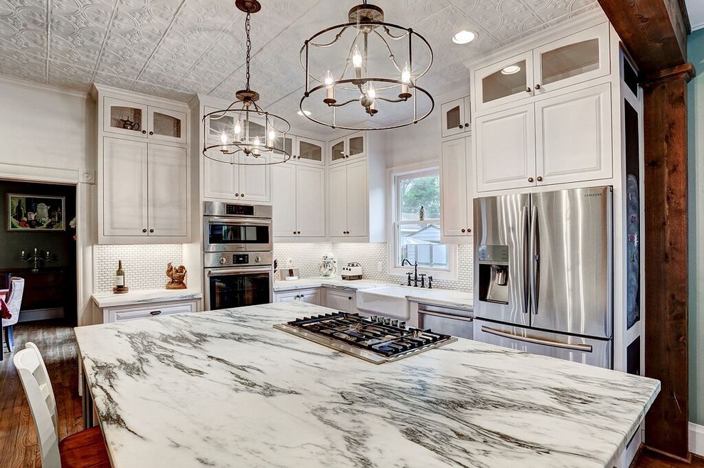 Photo of a farmhouse u-shaped kitchen/diner in Houston with a belfast sink, raised-panel cabinets, white cabinets, marble worktops, white splashback, porcelain splashback, stainless steel appliances, medium hardwood flooring, an island and brown floors.