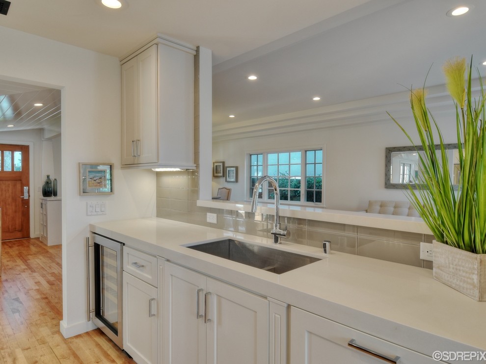 Small beach style galley open plan kitchen in San Diego with a belfast sink, shaker cabinets, white cabinets, composite countertops, white splashback, glass tiled splashback, stainless steel appliances and light hardwood flooring.