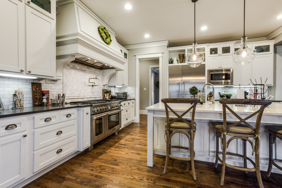 Elegant l-shaped medium tone wood floor open concept kitchen photo in Dallas with a farmhouse sink, shaker cabinets, white cabinets, quartz countertops, white backsplash, stone tile backsplash, stainless steel appliances and an island