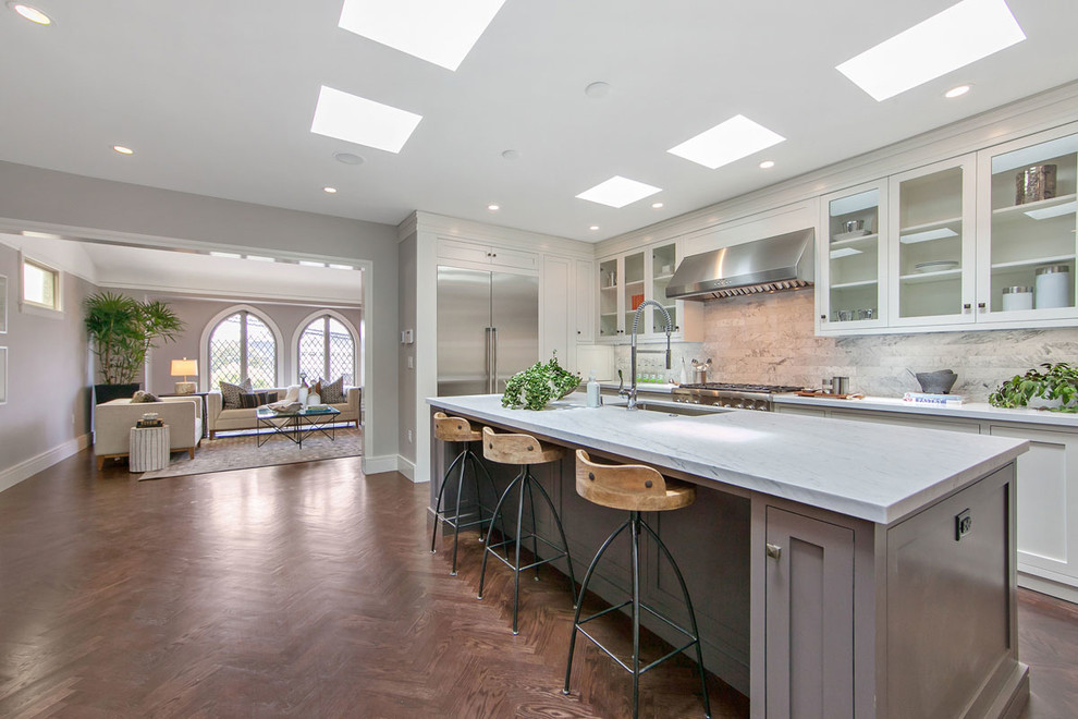 Modern inredning av ett mellanstort kök, med mörkt trägolv, en köksö, en undermonterad diskho, luckor med glaspanel, vita skåp, bänkskiva i kvarts, grått stänkskydd, stänkskydd i marmor, rostfria vitvaror och brunt golv