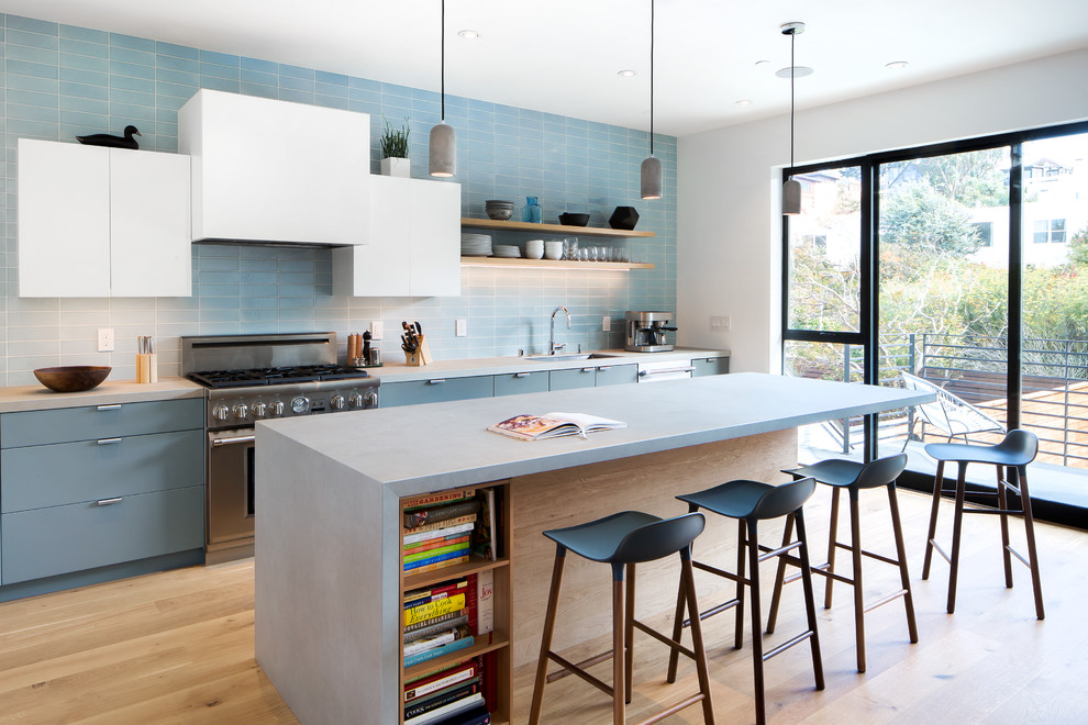 Modelo de cocina contemporánea con armarios con paneles lisos, puertas de armario azules, encimera de cemento, salpicadero azul, electrodomésticos de acero inoxidable, suelo de madera clara y una isla
