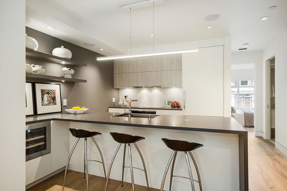 Photo of a large modern galley kitchen in San Francisco with a submerged sink, flat-panel cabinets, light wood cabinets, composite countertops, white splashback, glass sheet splashback, stainless steel appliances, medium hardwood flooring, a breakfast bar and brown floors.