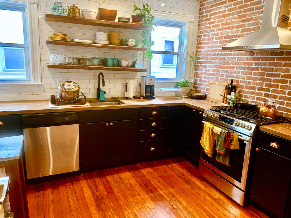 Inspiration for a mid-sized industrial u-shaped dark wood floor and brown floor enclosed kitchen remodel with a single-bowl sink, flat-panel cabinets, black cabinets, wood countertops, white backsplash, subway tile backsplash, stainless steel appliances, no island and brown countertops
