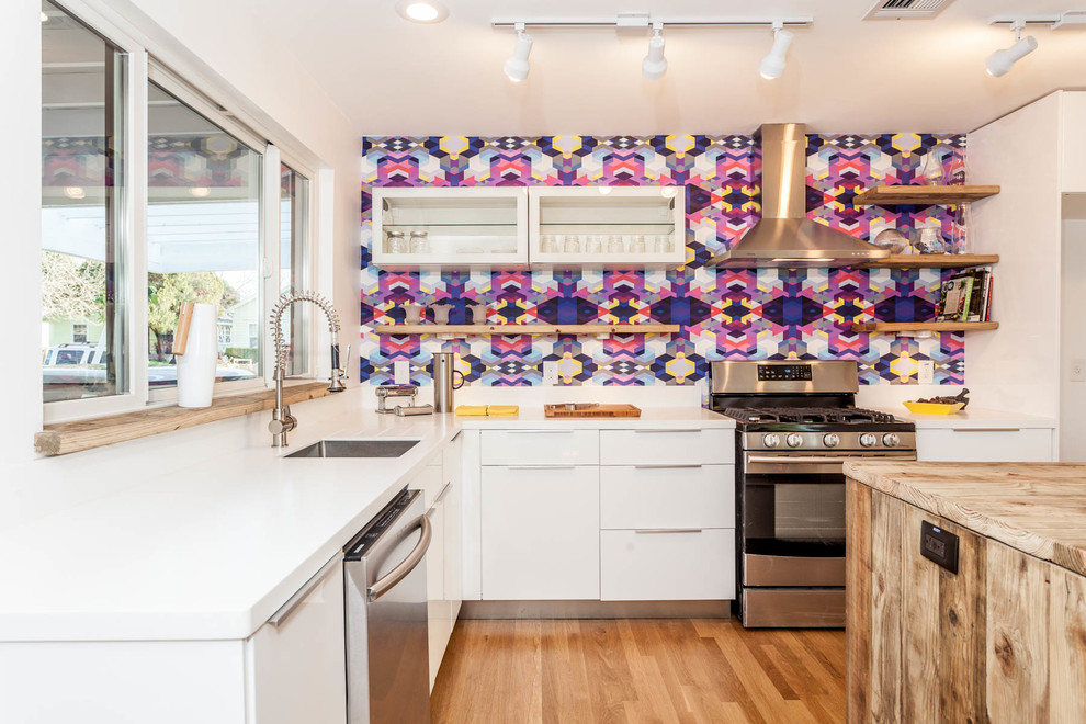 Foto på ett mellanstort minimalistiskt kök, med släta luckor, vita skåp, flerfärgad stänkskydd, rostfria vitvaror, ljust trägolv och en köksö