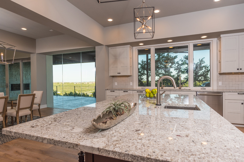 This is an example of a large contemporary l-shaped open plan kitchen in Austin with a double-bowl sink, shaker cabinets, white cabinets, white splashback, metro tiled splashback, stainless steel appliances, light hardwood flooring, an island and granite worktops.