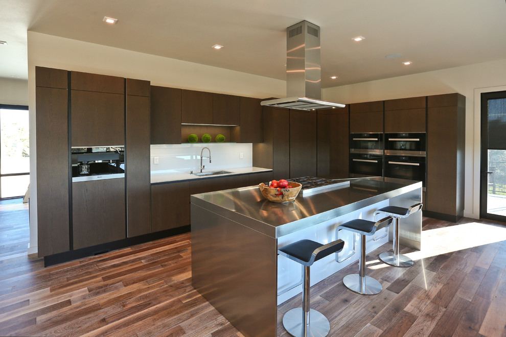Medium sized contemporary l-shaped kitchen/diner in Other with a submerged sink, flat-panel cabinets, medium wood cabinets, glass worktops, white splashback, glass sheet splashback, integrated appliances, medium hardwood flooring and an island.
