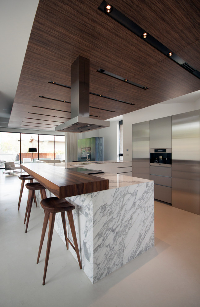 Modern inredning av ett kök, med en enkel diskho, släta luckor, skåp i rostfritt stål, marmorbänkskiva, rostfria vitvaror, betonggolv och en köksö