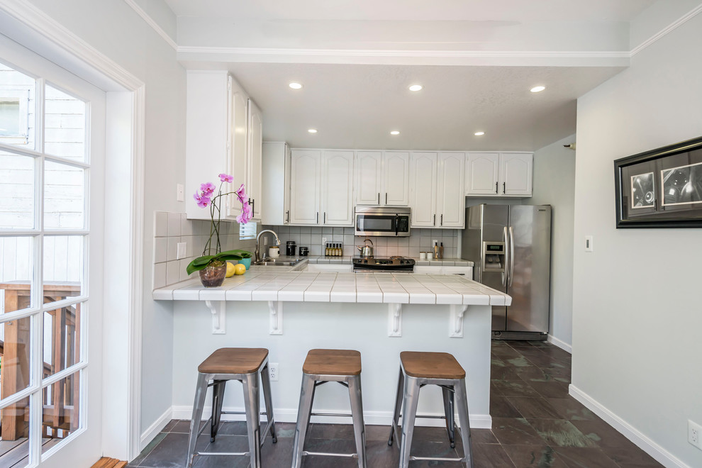 Example of a small transitional u-shaped ceramic tile and brown floor eat-in kitchen design in San Francisco with a double-bowl sink, raised-panel cabinets, white cabinets, tile countertops, white backsplash, ceramic backsplash, stainless steel appliances and a peninsula
