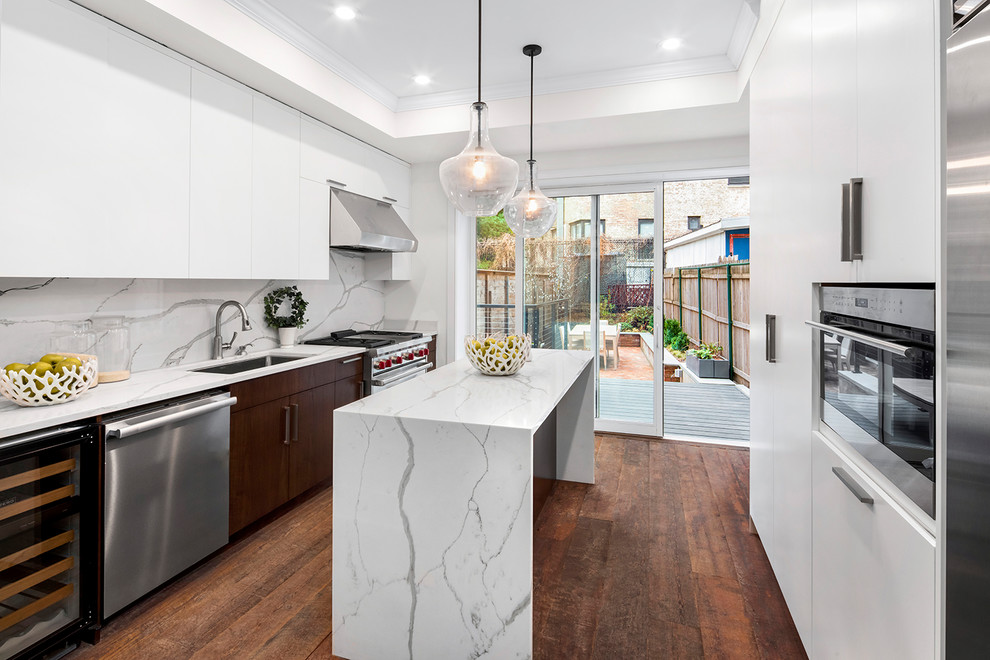 Traditional galley enclosed kitchen in New York with a single-bowl sink, flat-panel cabinets, white cabinets, white splashback, stainless steel appliances, dark hardwood flooring, an island and brown floors.