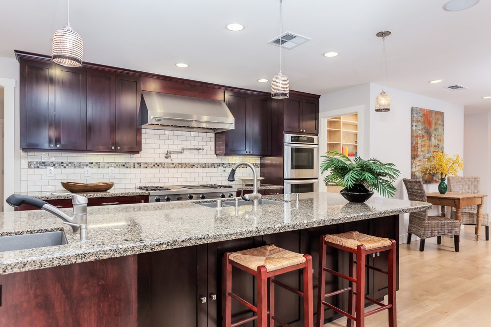 Photo of a large traditional single-wall kitchen/diner in San Francisco with a double-bowl sink, shaker cabinets, dark wood cabinets, engineered stone countertops, white splashback, metro tiled splashback, stainless steel appliances, light hardwood flooring and an island.