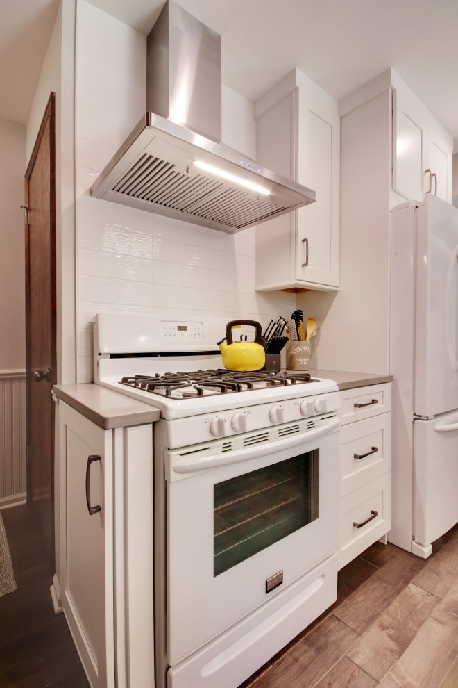This is an example of a small traditional u-shaped kitchen/diner in Grand Rapids with a submerged sink, recessed-panel cabinets, white cabinets, engineered stone countertops, white splashback, metro tiled splashback, white appliances, medium hardwood flooring, a breakfast bar, brown floors and grey worktops.