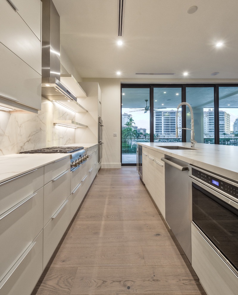 Large minimalist galley light wood floor and brown floor open concept kitchen photo in Miami with a drop-in sink, flat-panel cabinets, white cabinets, concrete countertops, white backsplash, porcelain backsplash, stainless steel appliances and an island