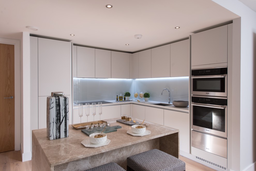 Contemporary l-shaped kitchen in London with a submerged sink, flat-panel cabinets, grey cabinets, grey splashback, glass sheet splashback, stainless steel appliances, light hardwood flooring, an island and white worktops.