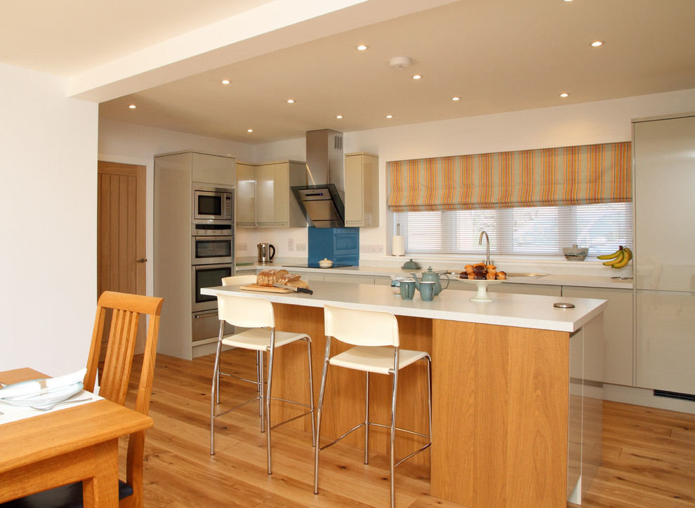 Offene, Mittelgroße Moderne Küche in L-Form mit flächenbündigen Schrankfronten, beigen Schränken, Küchenrückwand in Blau, Glasrückwand, Küchengeräten aus Edelstahl, braunem Holzboden und Kücheninsel in Kent