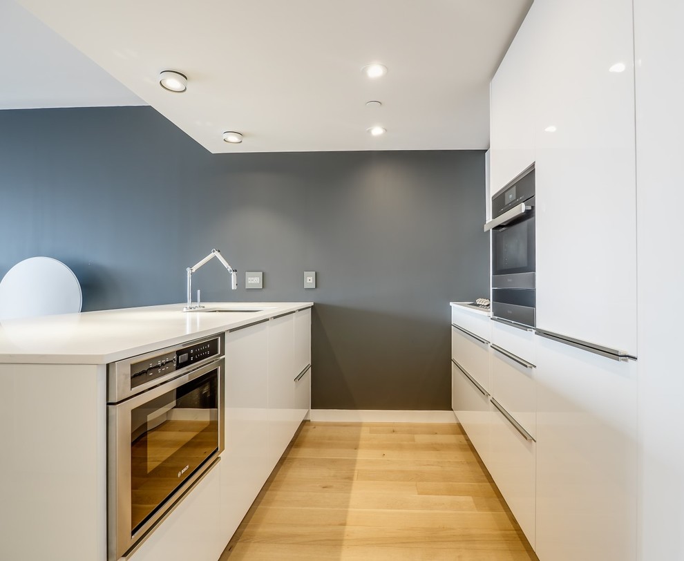 Mid-sized minimalist galley light wood floor and beige floor eat-in kitchen photo in New York with an undermount sink, flat-panel cabinets, white cabinets, white backsplash, stone slab backsplash, stainless steel appliances, a peninsula, quartz countertops and white countertops