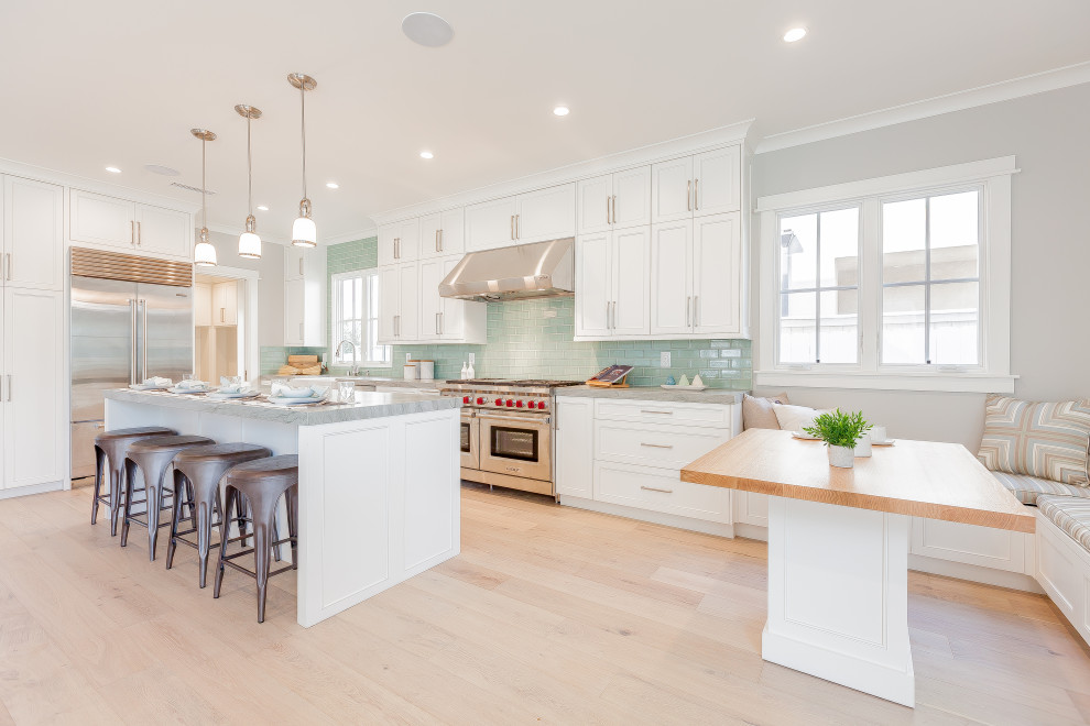 Design ideas for a large classic l-shaped kitchen/diner in San Diego with a belfast sink, shaker cabinets, white cabinets, green splashback, metro tiled splashback, stainless steel appliances, light hardwood flooring, an island, beige floors and grey worktops.
