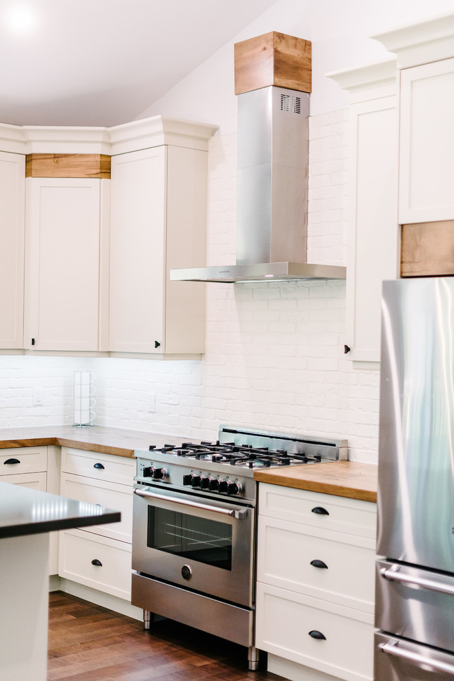 Example of a mid-sized minimalist l-shaped eat-in kitchen design with a farmhouse sink, shaker cabinets, white cabinets, wood countertops, white backsplash, brick backsplash, stainless steel appliances, an island and beige countertops