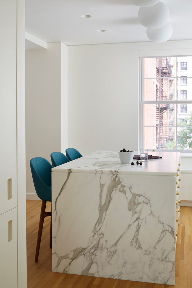 This is an example of a medium sized modern l-shaped kitchen/diner in New York with a submerged sink, flat-panel cabinets, white cabinets, marble worktops, grey splashback, ceramic splashback, integrated appliances, an island and light hardwood flooring.