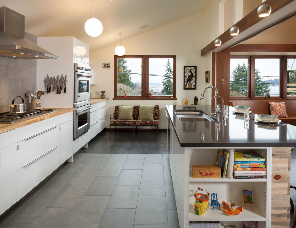 Example of a mid-sized trendy galley porcelain tile and gray floor open concept kitchen design in Seattle with an undermount sink, wood countertops, gray backsplash, stone tile backsplash, stainless steel appliances, an island, flat-panel cabinets and white cabinets