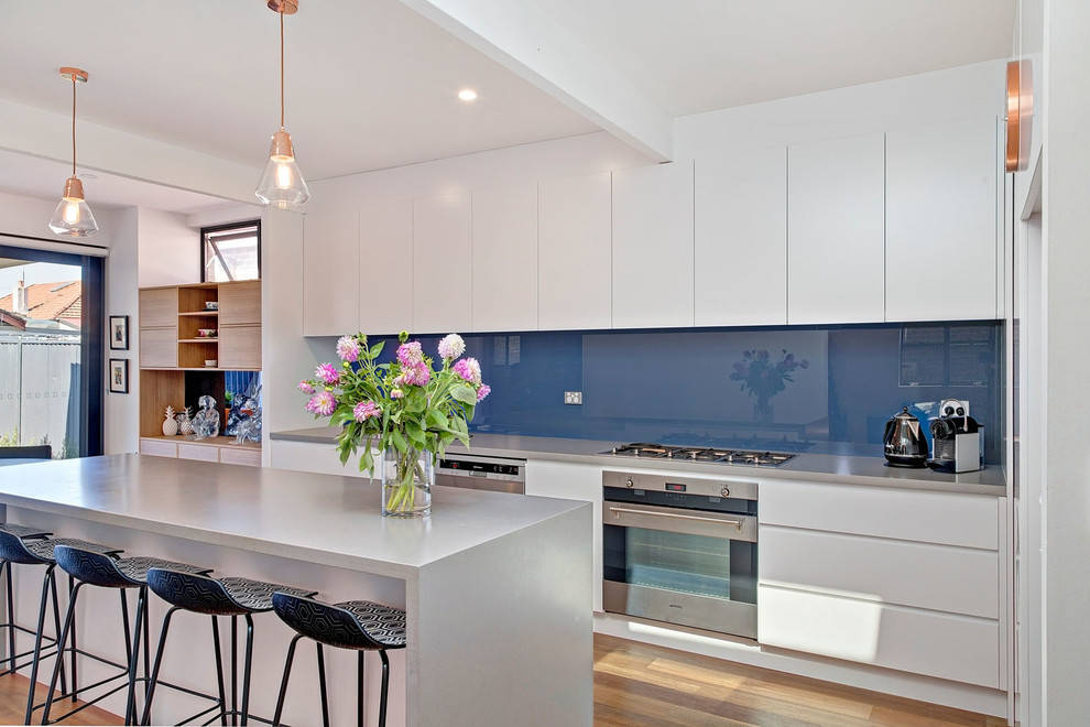 Moderne Küche mit flächenbündigen Schrankfronten, weißen Schränken, Küchenrückwand in Blau, Küchengeräten aus Edelstahl, braunem Holzboden und Kücheninsel in Sydney