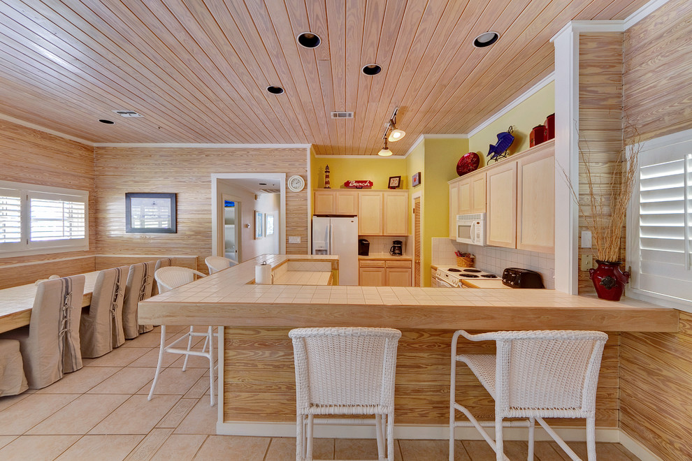 На фото: п-образная кухня в морском стиле с обеденным столом, фасадами с утопленной филенкой, светлыми деревянными фасадами, столешницей из плитки, бежевым фартуком, белой техникой и бежевой столешницей с