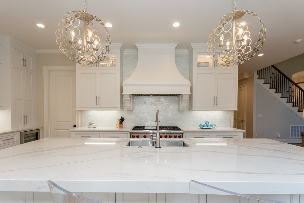 Bild på ett vit vitt kök och matrum, med en dubbel diskho, luckor med profilerade fronter, vita skåp, bänkskiva i kvarts, rostfria vitvaror, ljust trägolv och en köksö