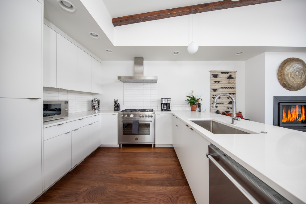 Foto på ett funkis vit u-kök, med en undermonterad diskho, släta luckor, vita skåp, rostfria vitvaror, mörkt trägolv, en halv köksö och brunt golv