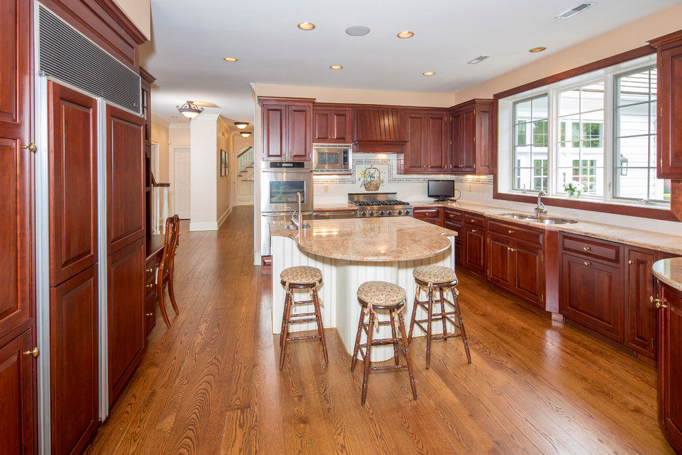 Inspiration for a large timeless light wood floor kitchen remodel in Philadelphia with a double-bowl sink, raised-panel cabinets, medium tone wood cabinets, granite countertops, white backsplash, ceramic backsplash, paneled appliances and an island