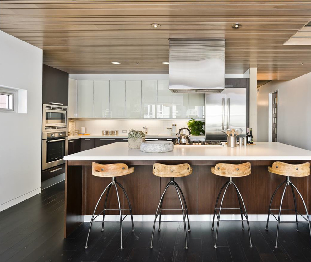 This is an example of a medium sized contemporary l-shaped open plan kitchen in San Francisco with stainless steel appliances, an integrated sink, flat-panel cabinets, dark wood cabinets, engineered stone countertops, white splashback, dark hardwood flooring and an island.
