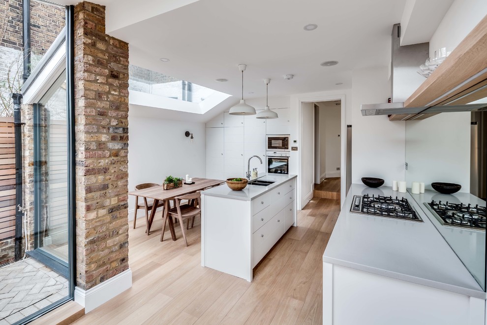 Zweizeilige Nordische Wohnküche mit Unterbauwaschbecken, flächenbündigen Schrankfronten, weißen Schränken, Rückwand aus Spiegelfliesen, hellem Holzboden und Kücheninsel in London