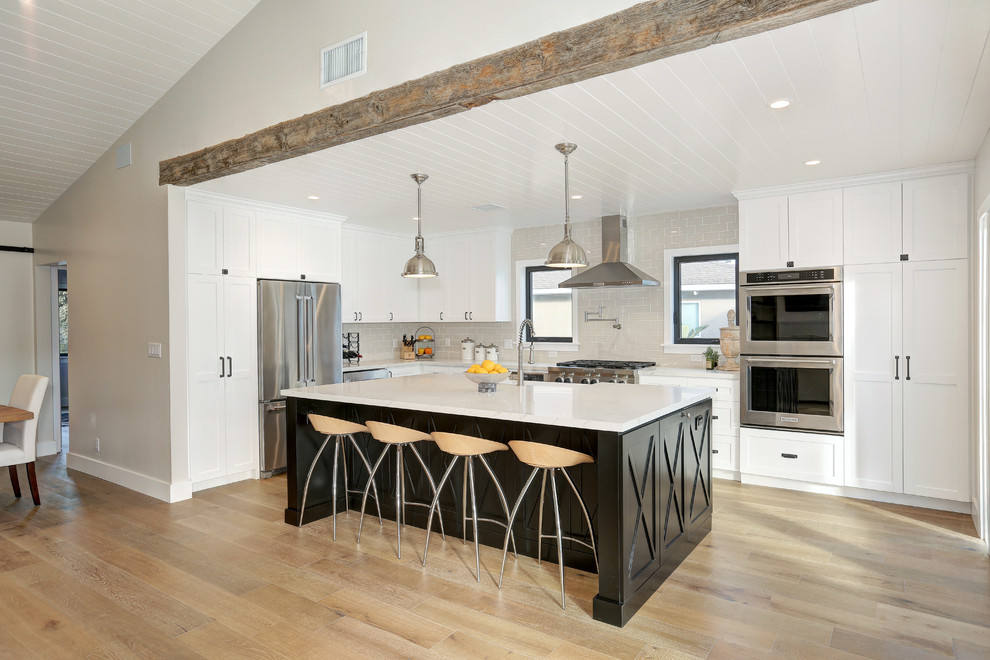 This is an example of a country u-shaped kitchen in Los Angeles with a belfast sink, shaker cabinets, white cabinets, grey splashback, stainless steel appliances, medium hardwood flooring, an island and brown floors.