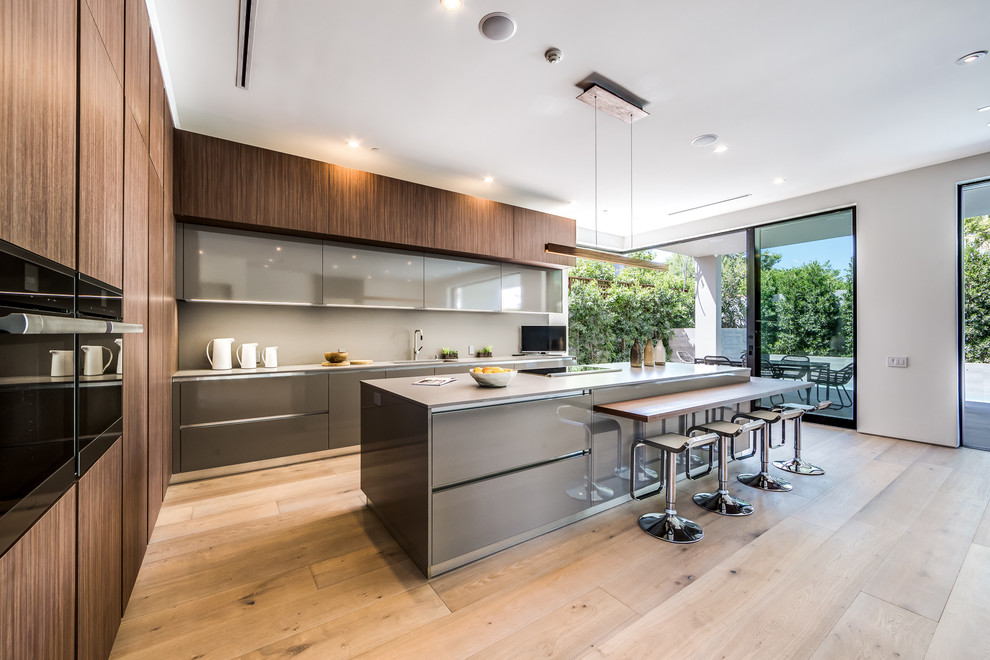 This is an example of a large modern open plan kitchen in Los Angeles with brown cabinets, granite worktops, grey splashback, light hardwood flooring, an island, flat-panel cabinets and stainless steel appliances.