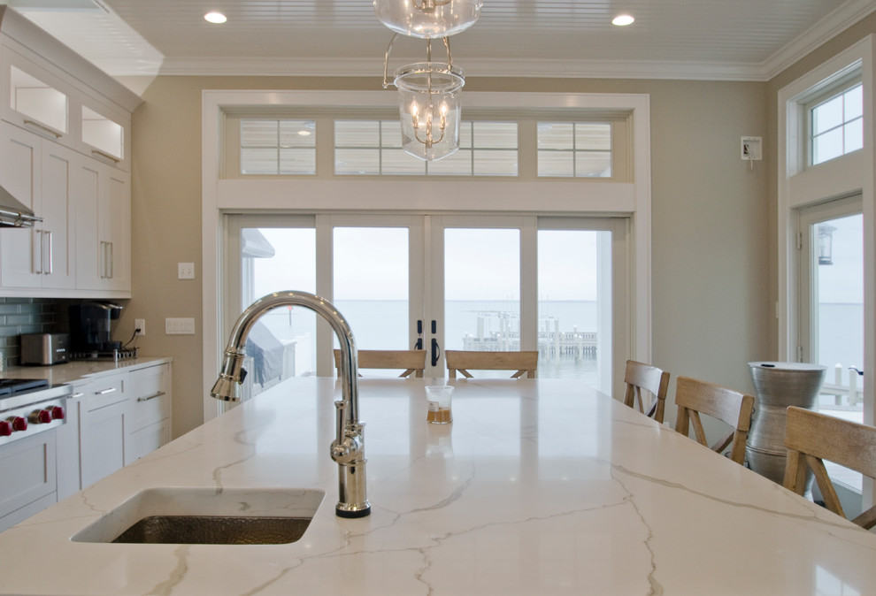 This is an example of a medium sized nautical l-shaped open plan kitchen in Charleston with a belfast sink, shaker cabinets, white cabinets, engineered stone countertops, grey splashback, glass tiled splashback, integrated appliances, light hardwood flooring, an island, brown floors and white worktops.