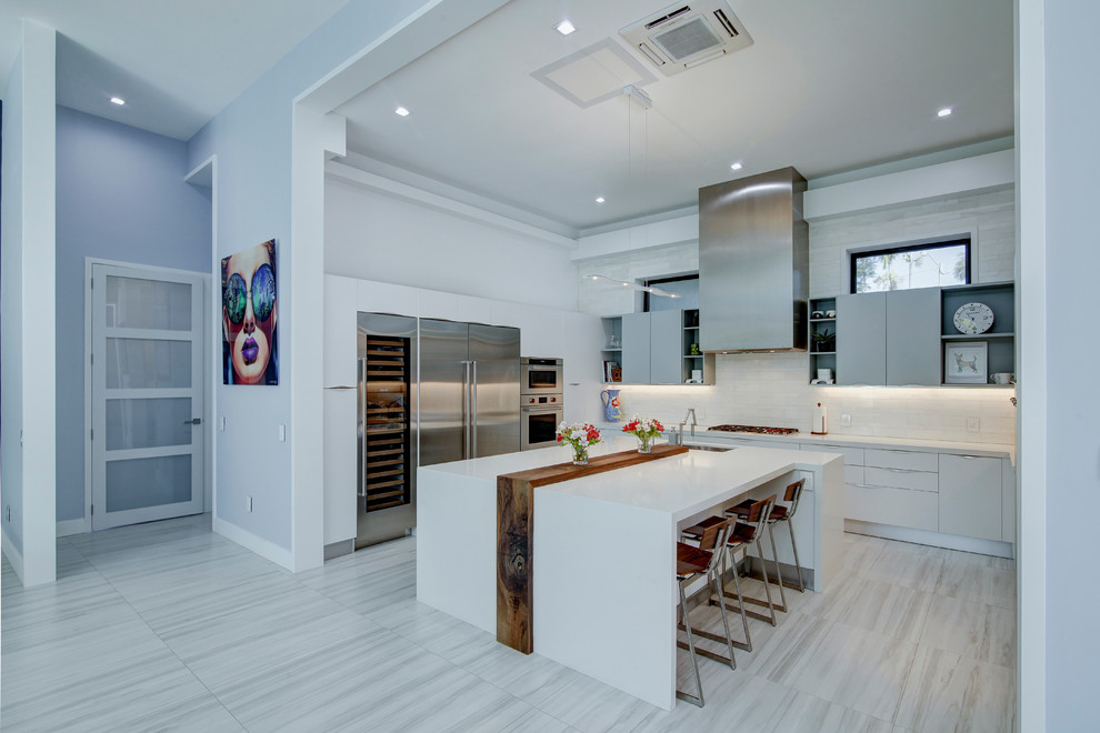Cette image montre une cuisine design en U avec un placard à porte plane, des portes de placard blanches, une crédence blanche, un électroménager en acier inoxydable, îlot et un sol gris.