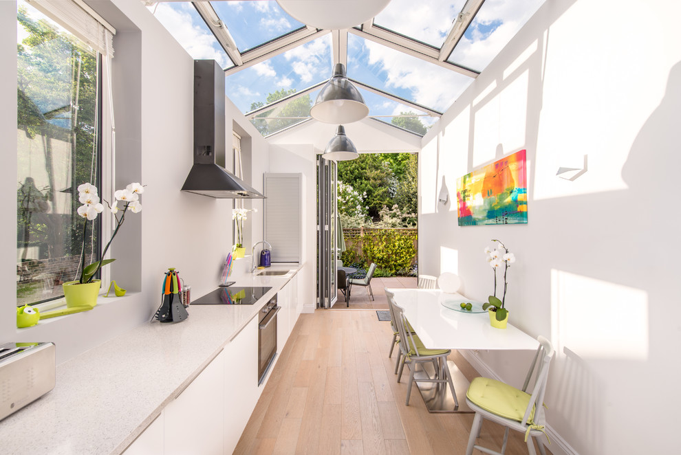 Inspiration for a medium sized contemporary galley kitchen/diner in London with a built-in sink, stainless steel appliances, light hardwood flooring, an island, flat-panel cabinets and white cabinets.