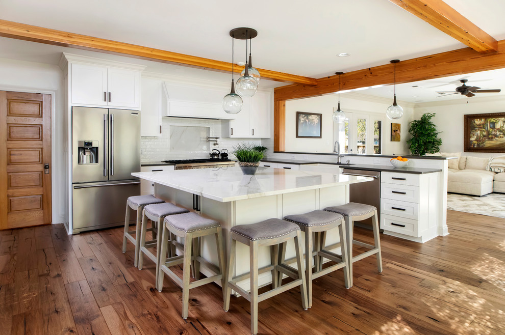 Photo of a large farmhouse l-shaped open plan kitchen in New Orleans with white cabinets, quartz worktops, white splashback, ceramic splashback, stainless steel appliances, medium hardwood flooring, an island, brown floors, white worktops, a belfast sink and shaker cabinets.