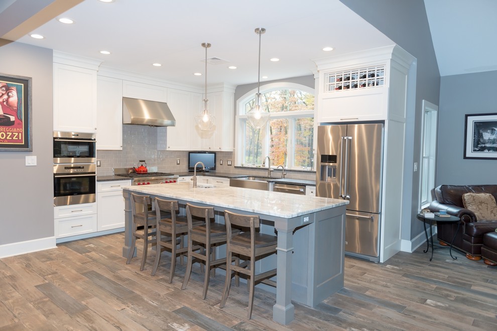 Inspiration for a mid-sized transitional l-shaped brown floor open concept kitchen remodel in New York with a farmhouse sink, shaker cabinets, white cabinets, marble countertops, gray backsplash, ceramic backsplash, stainless steel appliances, an island and white countertops