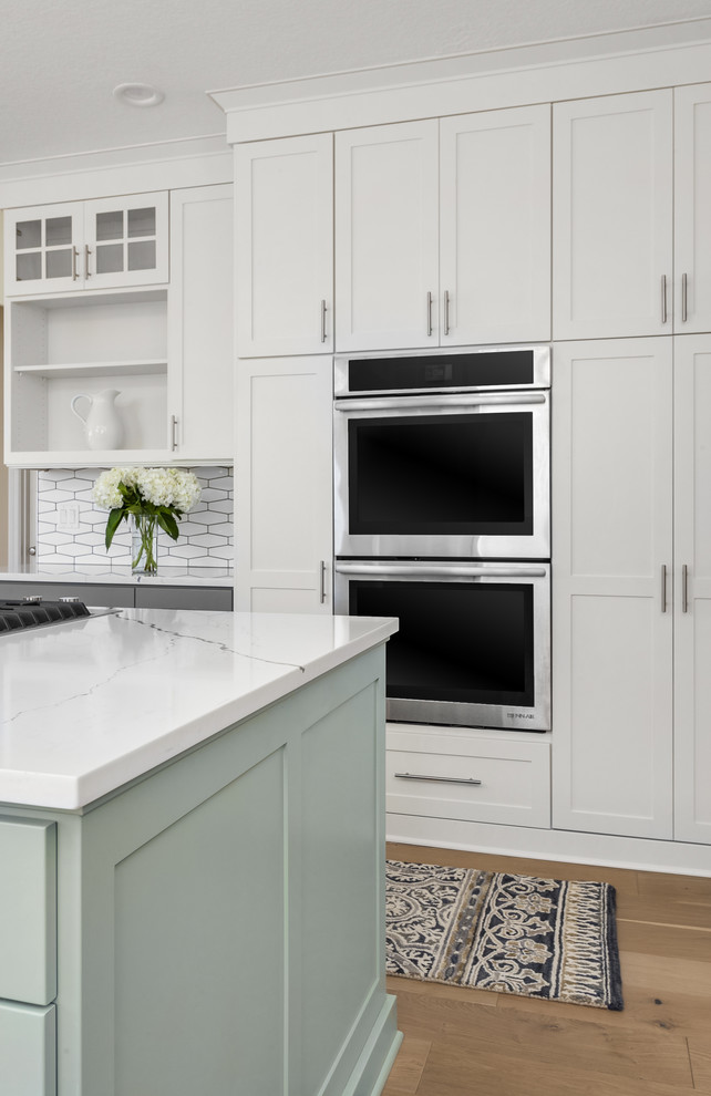 Foto på ett mellanstort lantligt vit kök, med en undermonterad diskho, skåp i shakerstil, grå skåp, bänkskiva i kvarts, vitt stänkskydd, stänkskydd i porslinskakel, rostfria vitvaror, mellanmörkt trägolv och en köksö