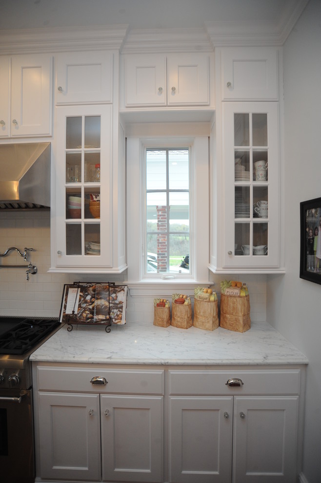 Example of a classic l-shaped enclosed kitchen design in Other with a farmhouse sink, glass-front cabinets, white cabinets, marble countertops, white backsplash, subway tile backsplash and stainless steel appliances