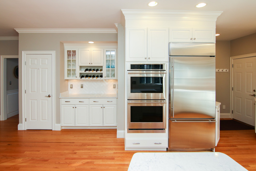 Bild på ett stort vintage vit vitt kök, med en undermonterad diskho, luckor med infälld panel, vita skåp, bänkskiva i kvartsit, vitt stänkskydd, stänkskydd i tunnelbanekakel, rostfria vitvaror, mellanmörkt trägolv, en halv köksö och brunt golv