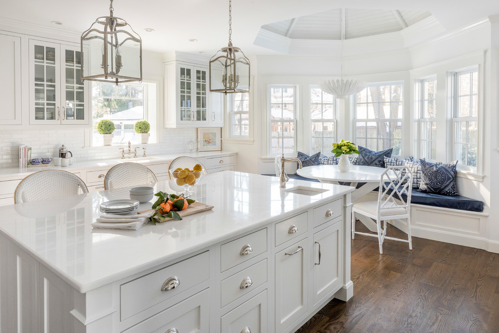 Inspiration for a large classic u-shaped open plan kitchen in Boston with an integrated sink, recessed-panel cabinets, white cabinets, composite countertops, white splashback, ceramic splashback, stainless steel appliances, dark hardwood flooring, an island, brown floors and white worktops.