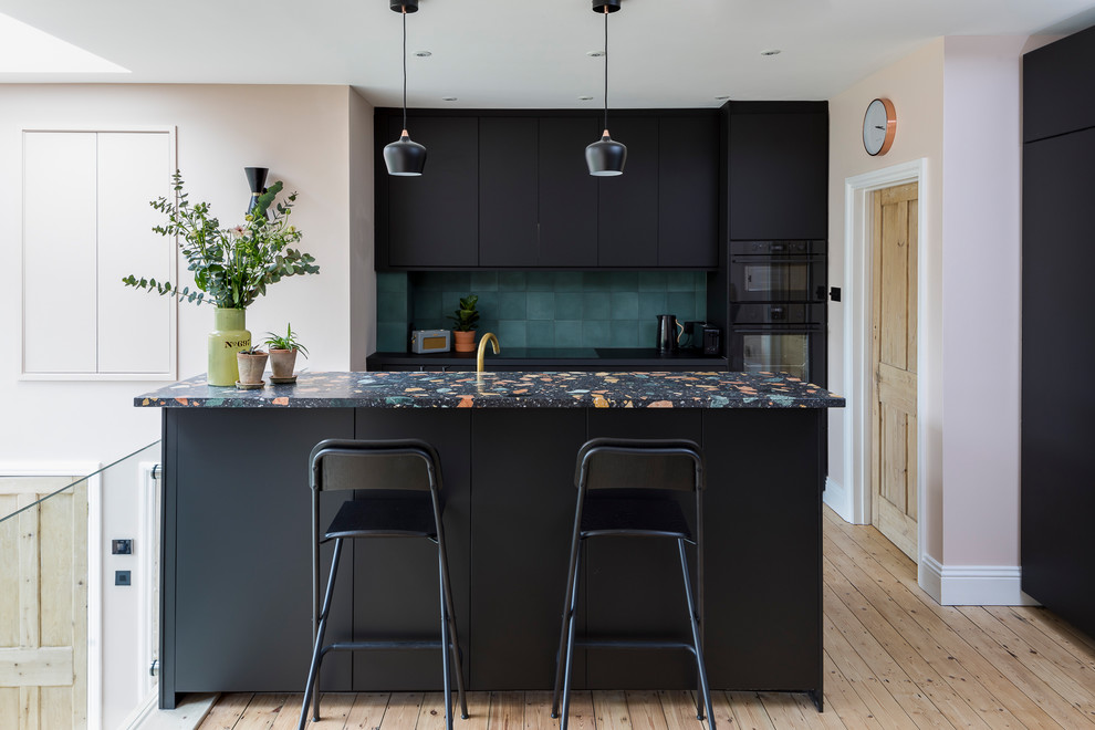 Nordische Küche mit flächenbündigen Schrankfronten, schwarzen Schränken, Küchenrückwand in Grün, schwarzen Elektrogeräten, hellem Holzboden, Kücheninsel, beigem Boden, bunter Arbeitsplatte und Arbeitsplatte aus Terrazzo in London